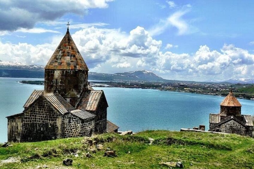 Une église à toit en flèche et un bâtiment plus petit surmonté d’un dôme, tous deux construits en pierre, surplombent un lac paisible, avec des montagnes visibles au loin. 2