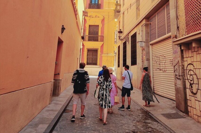 Una calle estrecha flanqueada por edificios, un grupo de personas caminando y una tienda cerrada con persiana. 1