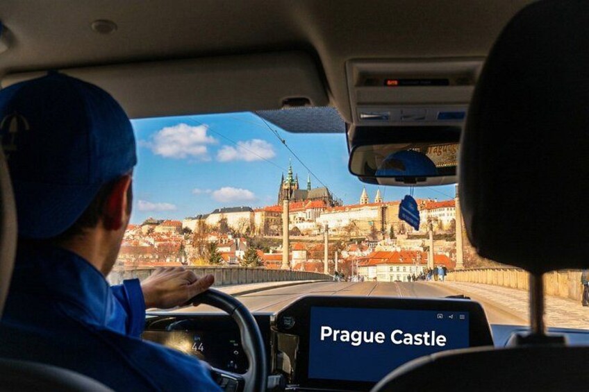 A driver in a blue uniform with a map application displaying 'Prague Castle' on the dashboard. 2