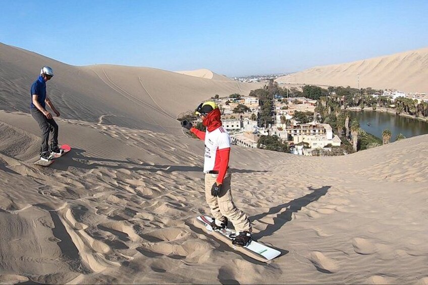 Twee mensen snowboarden op een zandduin, met een stadsgezicht op de achtergrond. 1