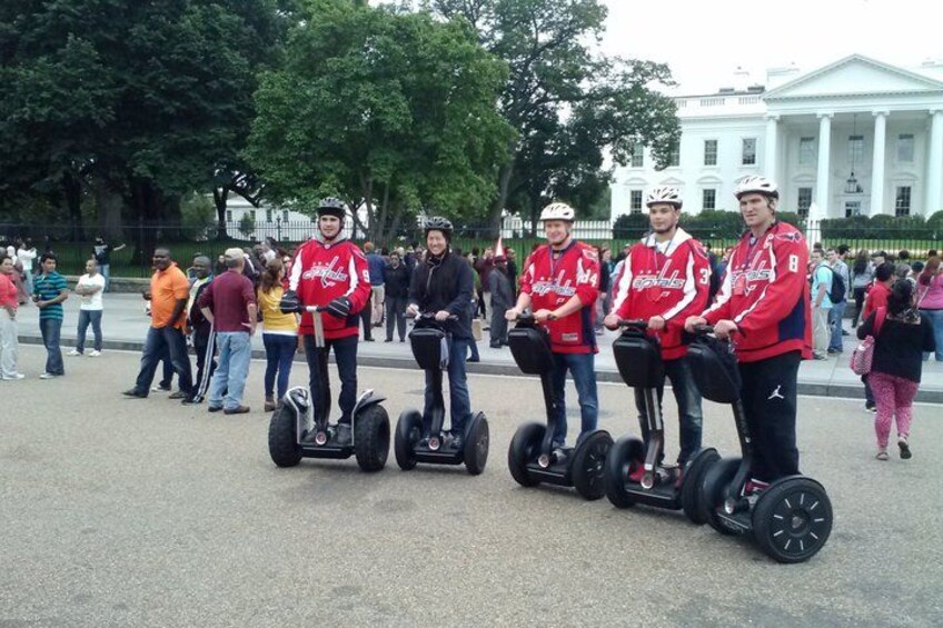 A group of people on segways in a public square with a large white building in the background. 2