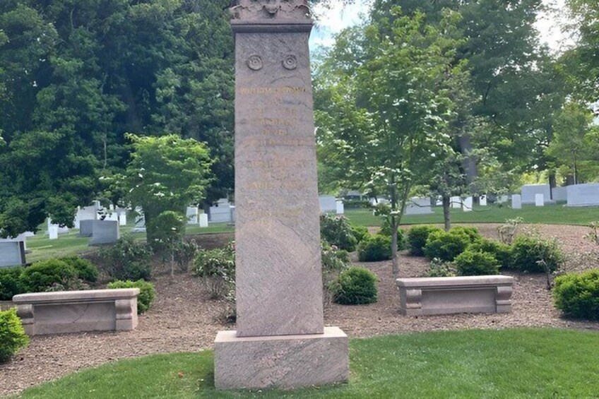 A stone monument with inscriptions, surrounded by a cemetery with headstones and trimmed bushes. 2