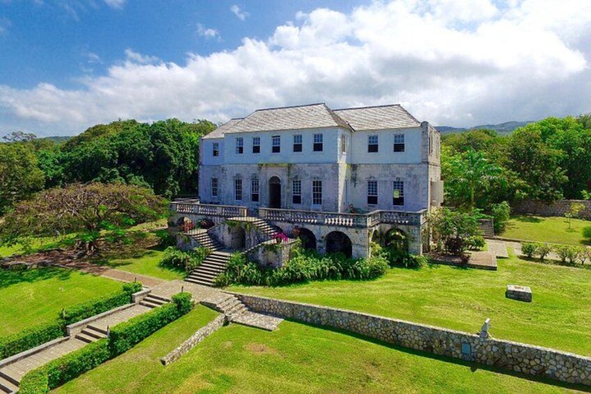 A large, white, two-story house with a stone staircase leading to a bridge, surrounded by lush greenery and a stone wall. 5