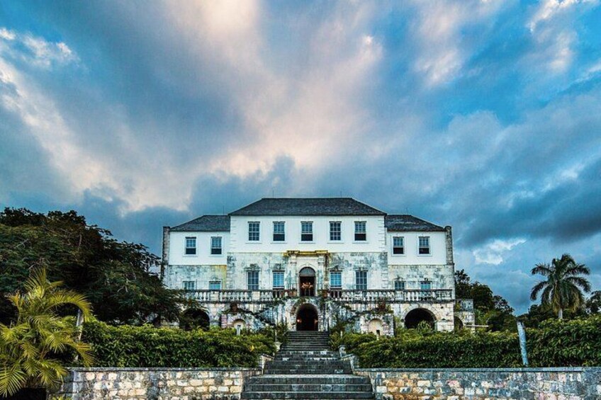 A large, two-story building with a central entrance, surrounded by lush greenery and palm trees. 2