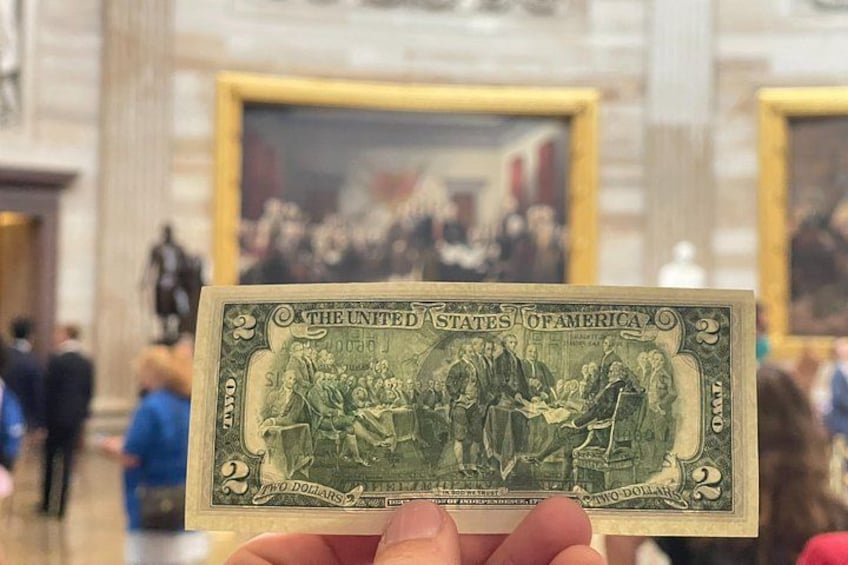 A hand holding a two-dollar bill with a historical scene on the front. 5