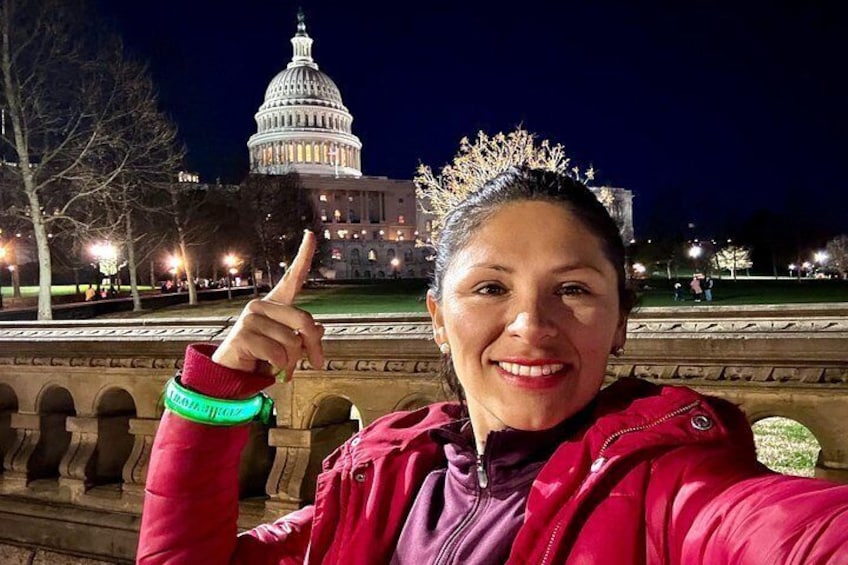 A person wearing a red jacket and a tiara, pointing upwards with a green wristband, with the Capitol building illuminated in the background. 3