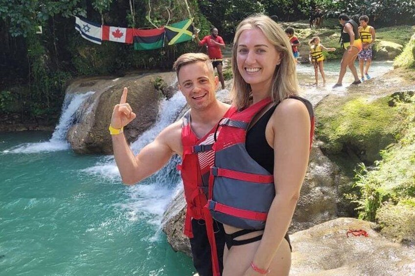 Un couple, vêtu d’un gilet de sauvetage rouge, se tient près d’une cascade, avec des drapeaux en arrière-plan. 2