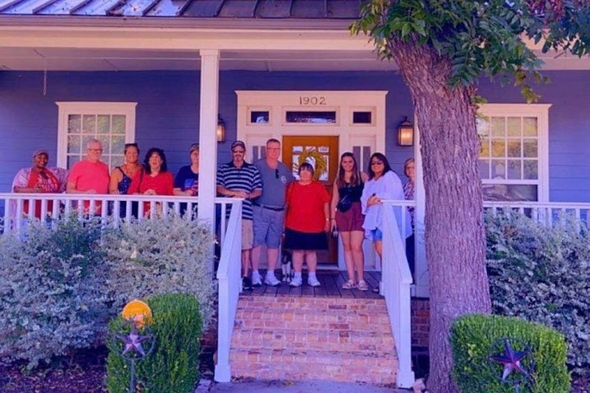 A group of people standing on a porch with a tree and a house number 1902. 5