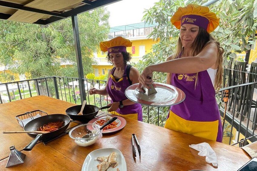 Two people cooking outdoors, wearing purple aprons and yellow hats, with a wooden table and cooking utensils. 2