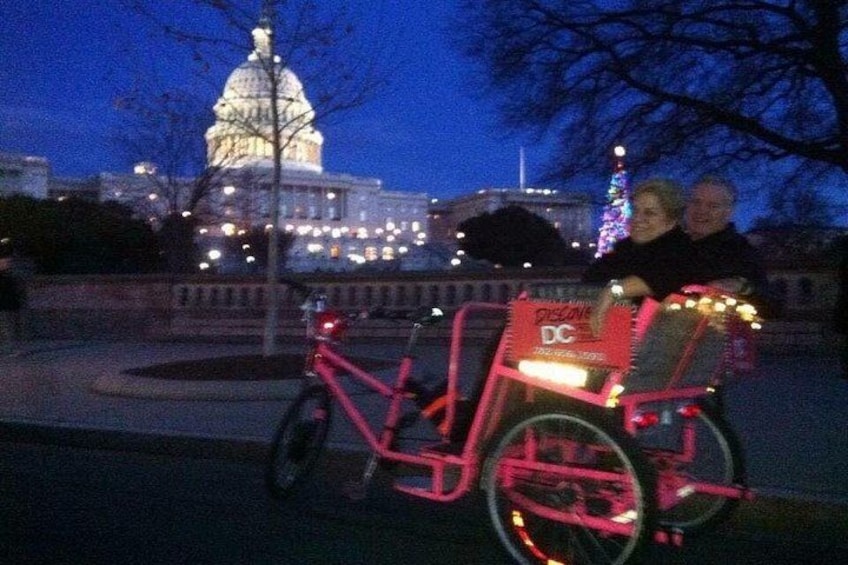 En rosa cykel med en DC-banner, en person framför och en byggnad med lampor i bakgrunden. 4