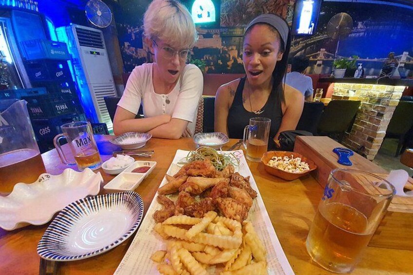 Two people at a restaurant table with a large plate of fried chicken and fries, a beer, and a sports bar in the background. 4