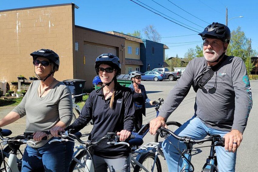 Three people on bicycles in an urban setting with buildings and parked cars in the background. 3