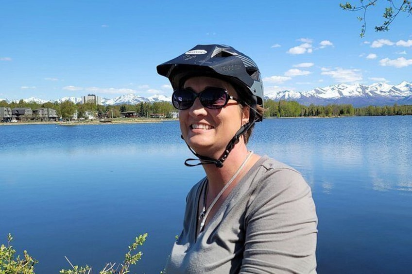 En person med hjelm og solbriller ved en rolig sø med bjerge i baggrunden. 2