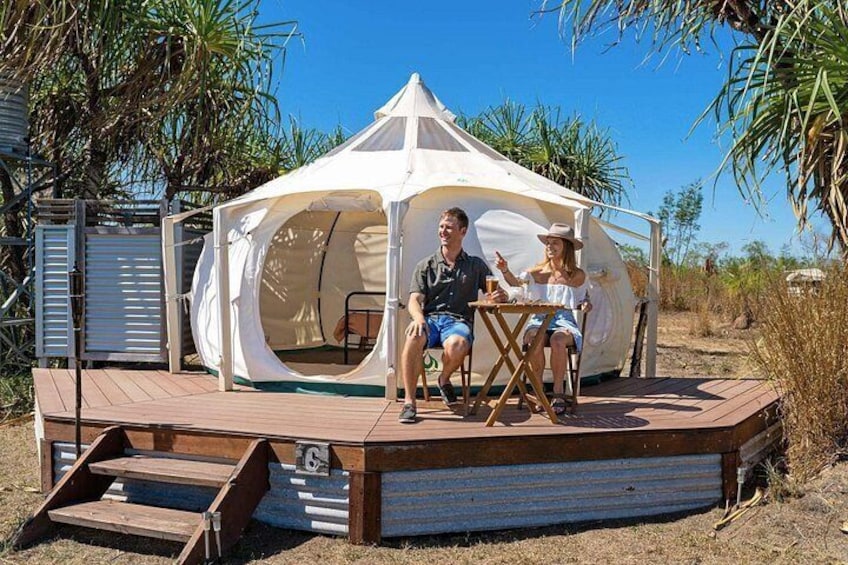 Ein Zelt mit einer Holzterrasse, zwei Personen sitzen an einem Tisch, im Hintergrund Palmen. 3