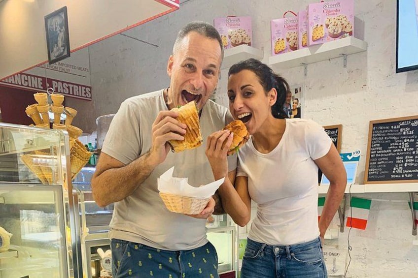 Two people eating sandwiches in a bakery with pastries and a menu board in the background. 1