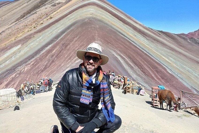 Una persona con chaqueta y sombrero negro, de pie frente a una colorida cordillera, con una multitud de fondo. 2