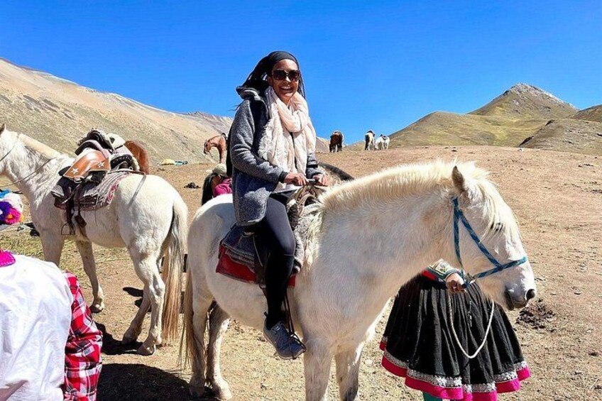 A person riding a white horse with a saddle and a black decorative cloth, in a mountainous area with other horses and riders in the background. 5