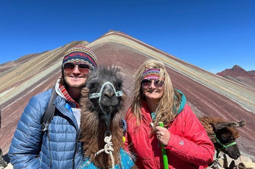 Eine Person in einer blauen Jacke, eine andere in einer roten Jacke, und ein Lama in einer gebirgigen Landschaft mit markanten roten und weißen Felsformationen. 5