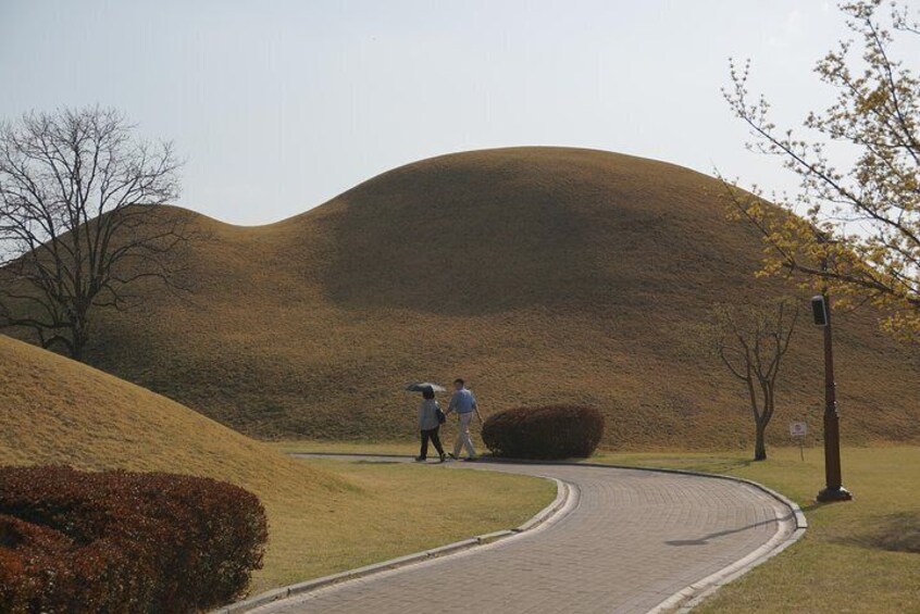 A curved pathway with two people walking, a large hill, and a bushy area. 1