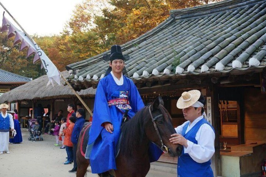 A person in traditional blue attire riding a horse, with a person in a hat interacting with the horse. 1