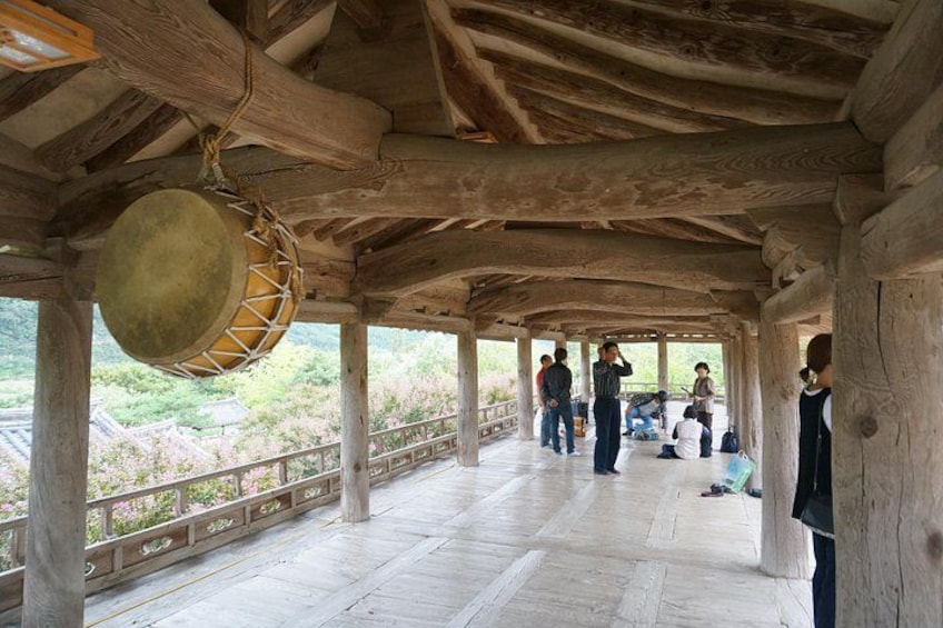 A wooden pavilion with a drum hanging from the ceiling, people sitting and standing on the floor, and a view of greenery outside. 4