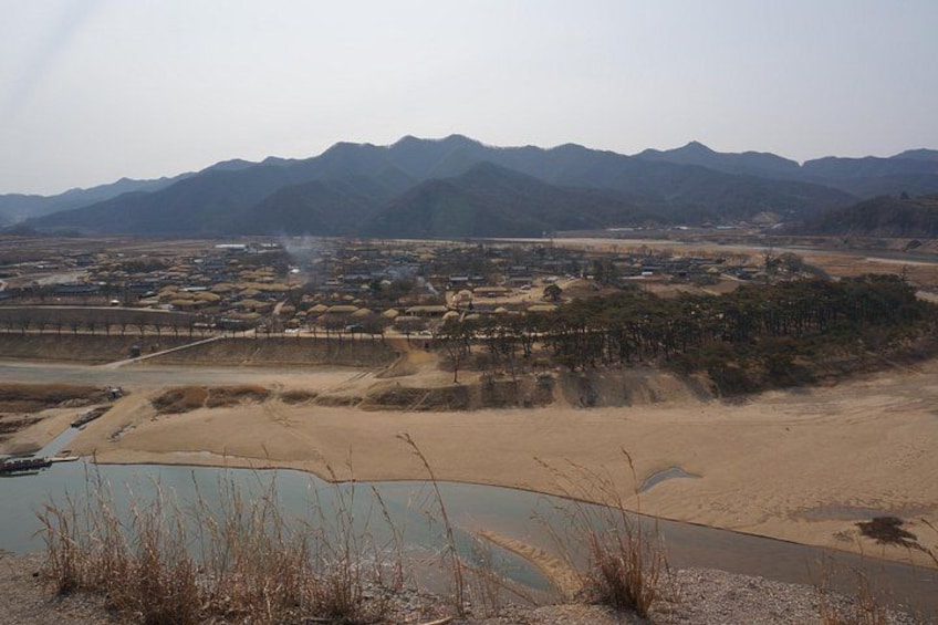 A dry riverbed with sparse vegetation, a distant settlement, and mountains in the background. 3