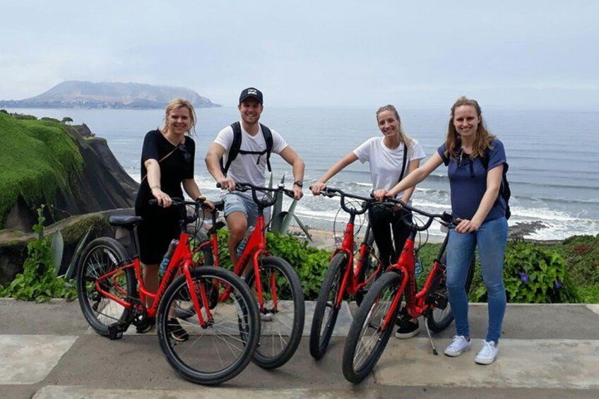 Four people on bicycles by the ocean with a cliff and greenery in the background. 3