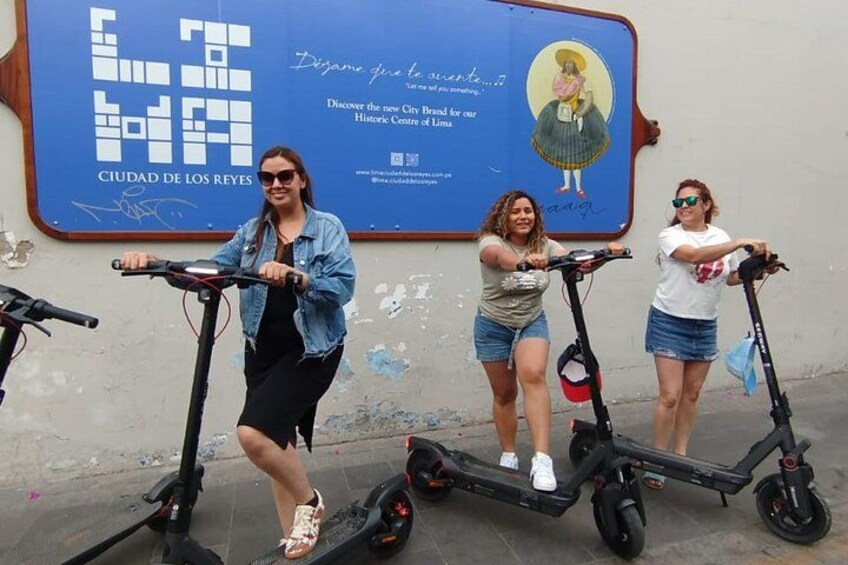 Three people riding electric scooters on a street with a blue advertisement in the background. 2