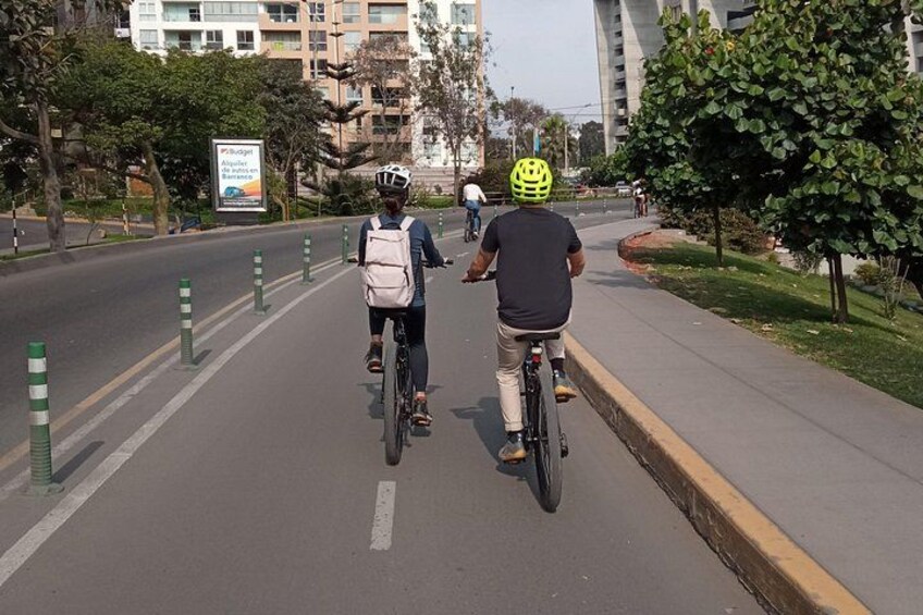 Two cyclists on a paved path with greenery and buildings in the background. 1