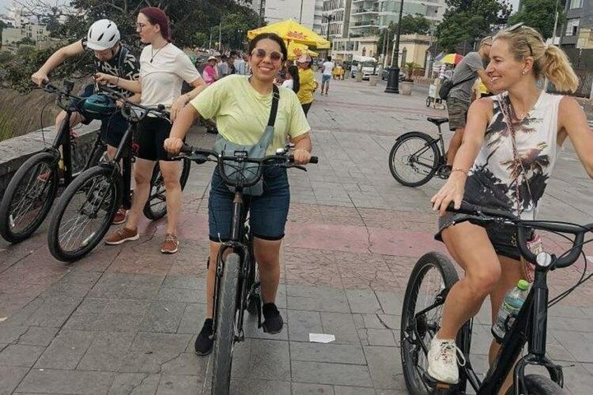 People riding bicycles on a paved path with buildings in the background. 4
