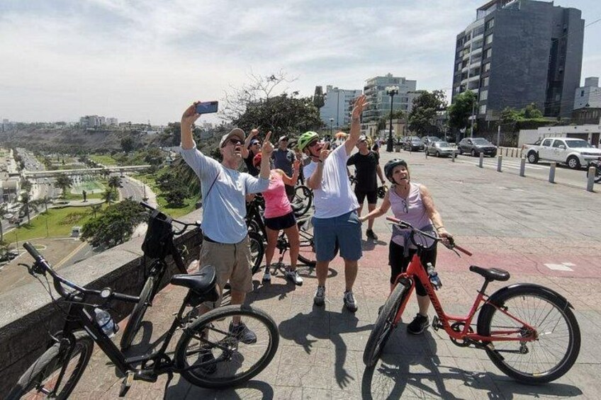 A group of people with bicycles, one person taking a selfie, and a cityscape in the background. 5