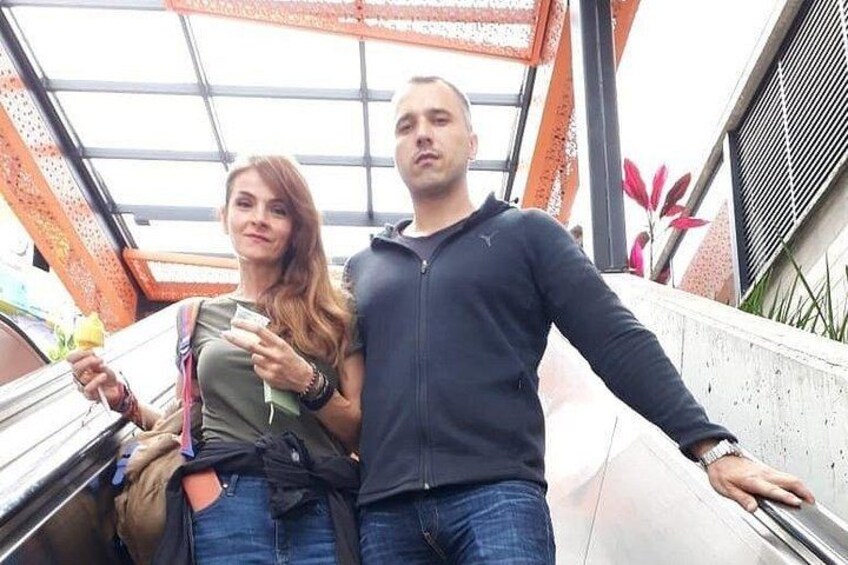 A man and a woman standing on a bridge with a metal railing, holding hands, and looking at the camera. 3