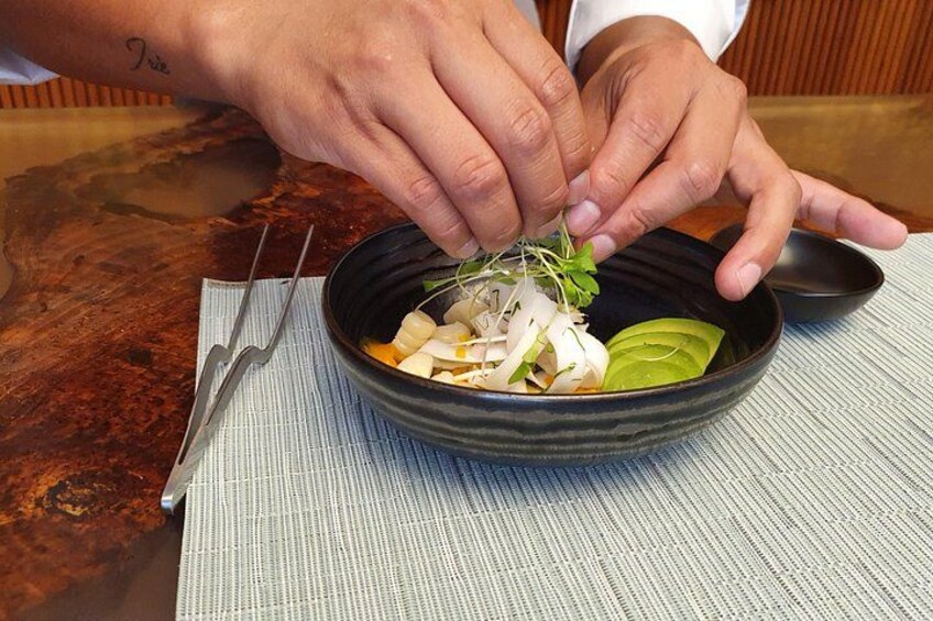 A person preparing a dish with vegetables and herbs in a black bowl on a wooden table. 2