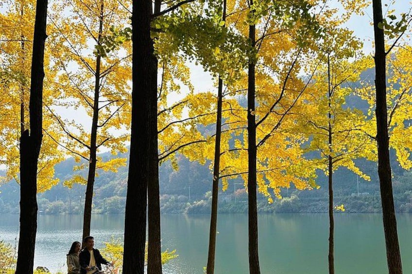 A lake surrounded by tall trees with yellow leaves, a couple sitting on a bench, and a mountainous landscape in the background. 2
