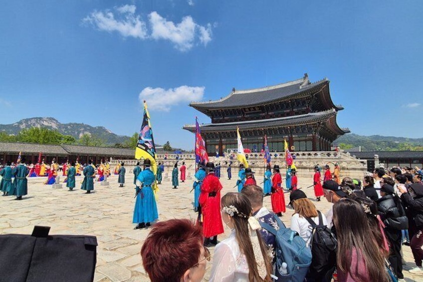 Una ceremonia tradicional coreana con participantes vestidos con vibrantes hanbok, un palacio histórico y un paisaje montañoso al fondo. 1