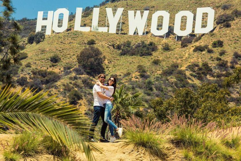 Una pareja abrazándose frente al cartel de Hollywood, con un paisaje montañoso de fondo. 1