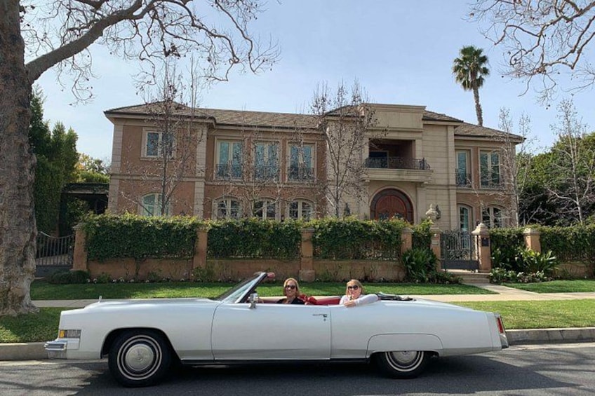 A white convertible car parked in front of a large, multi-story house with a well-maintained lawn and a palm tree. 3