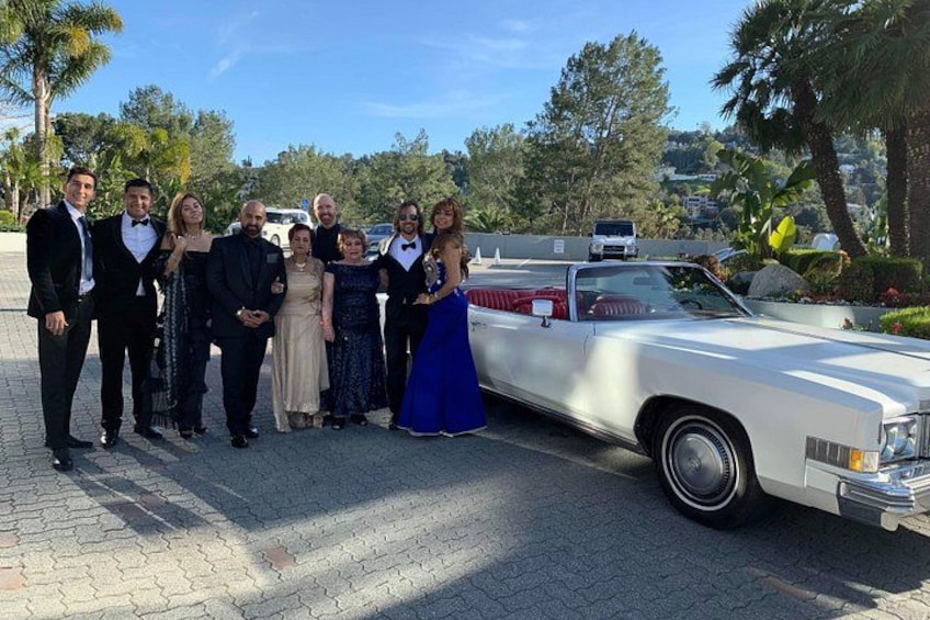A group of people posing in front of a white vintage car on a paved area with palm trees and a clear sky in the background. 4