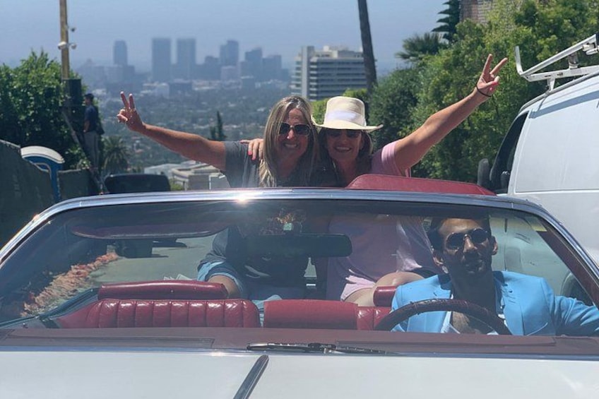 A vintage car with red interior, a cityscape in the background, and a person making a peace sign. 2