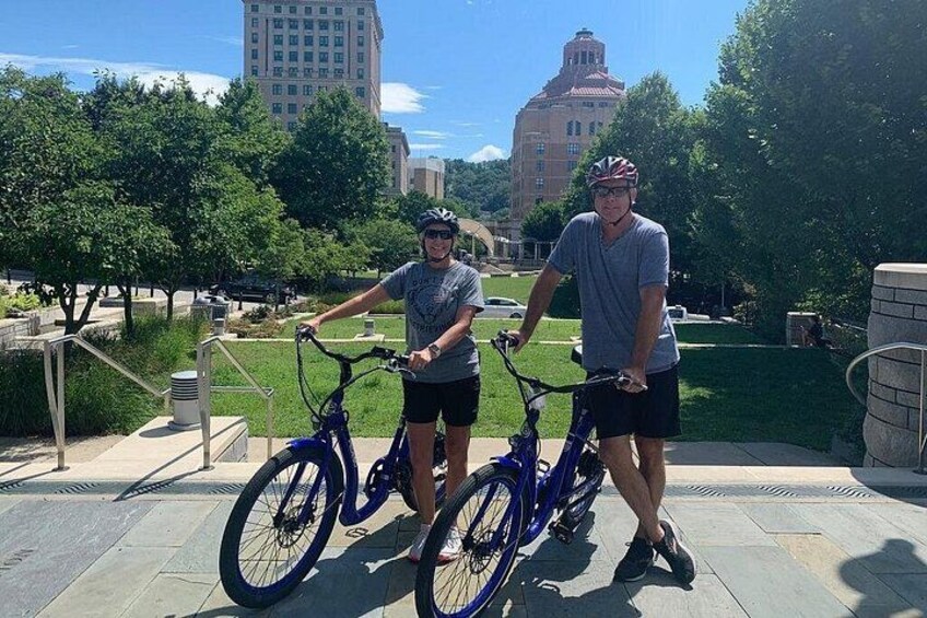 Dos personas de pie junto a bicicletas en un camino pavimentado, con árboles y edificios al fondo. 2