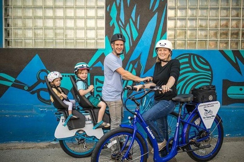 En person cykler på en blå cykel med et barn på en trehjulecykel og et andet barn på en løbehjælper foran en mural med abstrakte motiver. 3