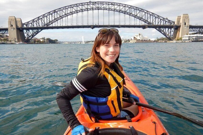 A person in a kayak with a life jacket, paddling on water with the Sydney Opera House and Sydney Harbour Bridge in the background. 1
