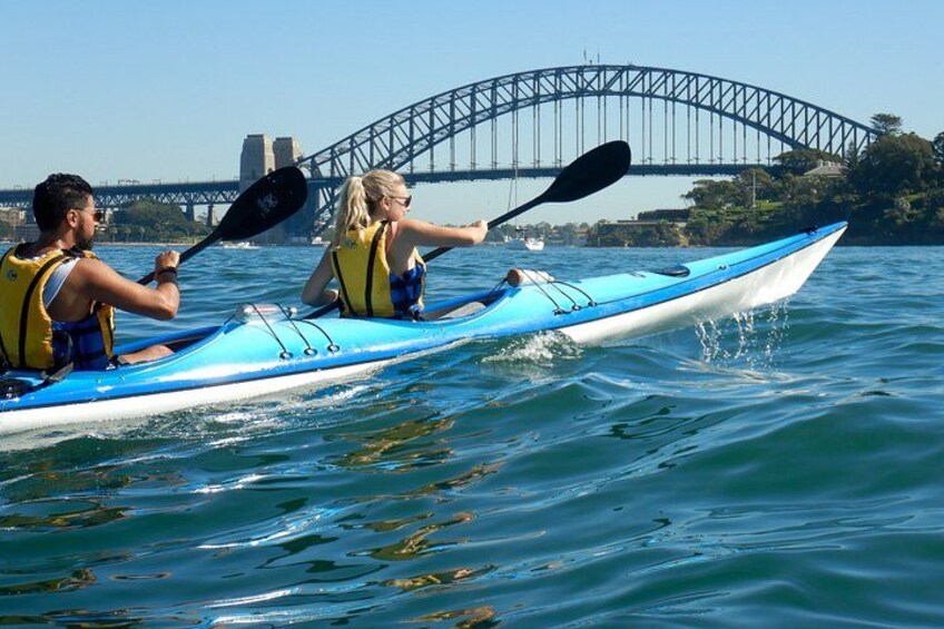 Two people kayaking on a river with a bridge in the background. 2