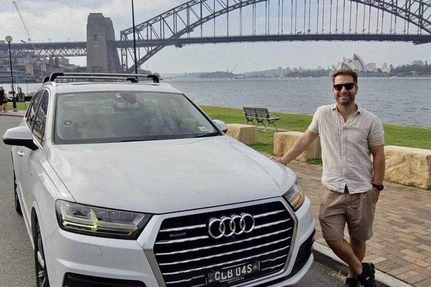 A white Audi SUV parked by a waterfront with a bridge and Sydney Opera House in the background. 3