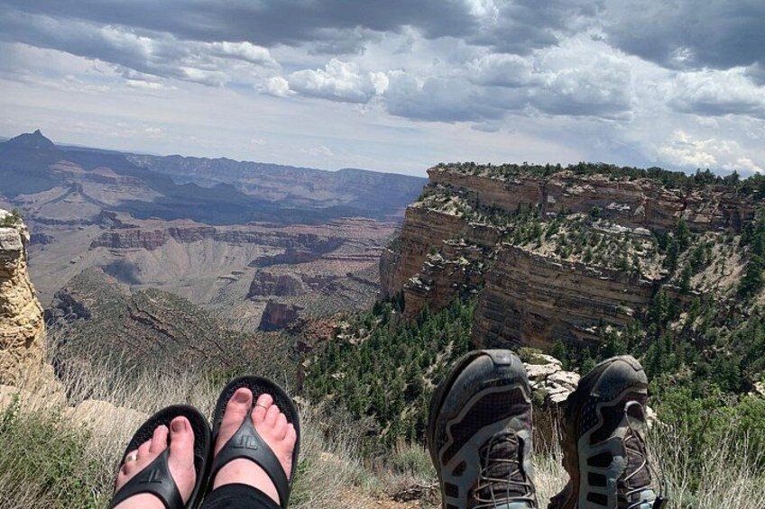 Les pieds d’une personne, chaussés d’une paire de chaussures de randonnée et d’une paire de sandales, surplombent un canyon caractérisé par des formations rocheuses en strates et une végétation clairsemée. 3