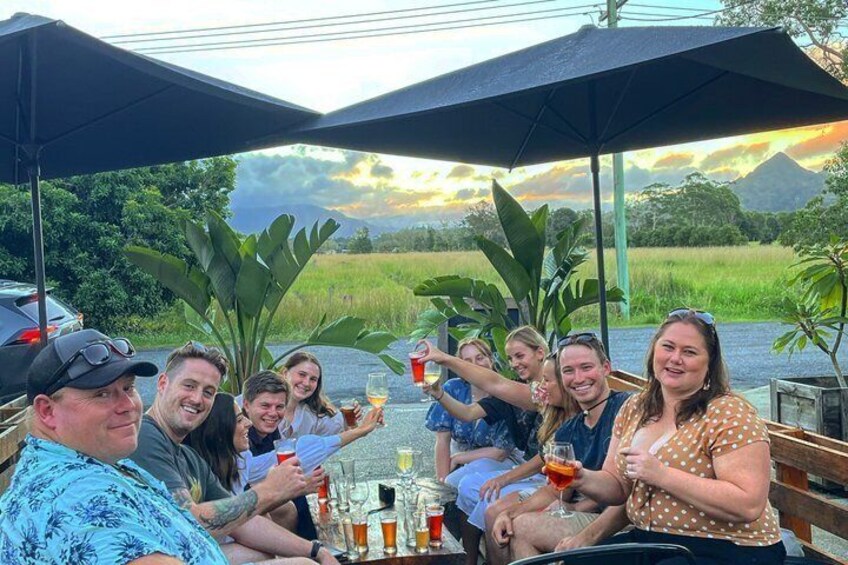 A group of people sitting at an outdoor table with drinks, surrounded by umbrellas and plants, with a scenic landscape in the background. 5