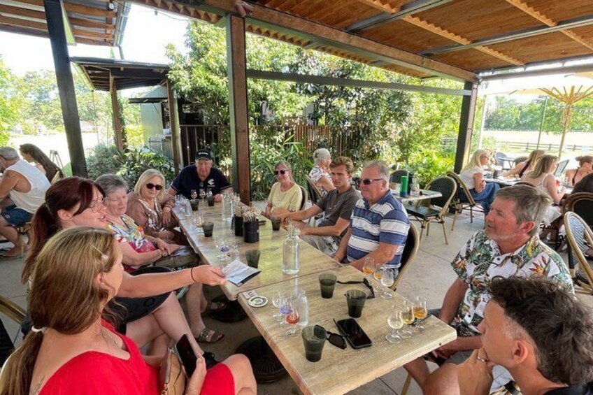A group of people seated at an outdoor table with drinks, under a covered area with greenery in the background. 3