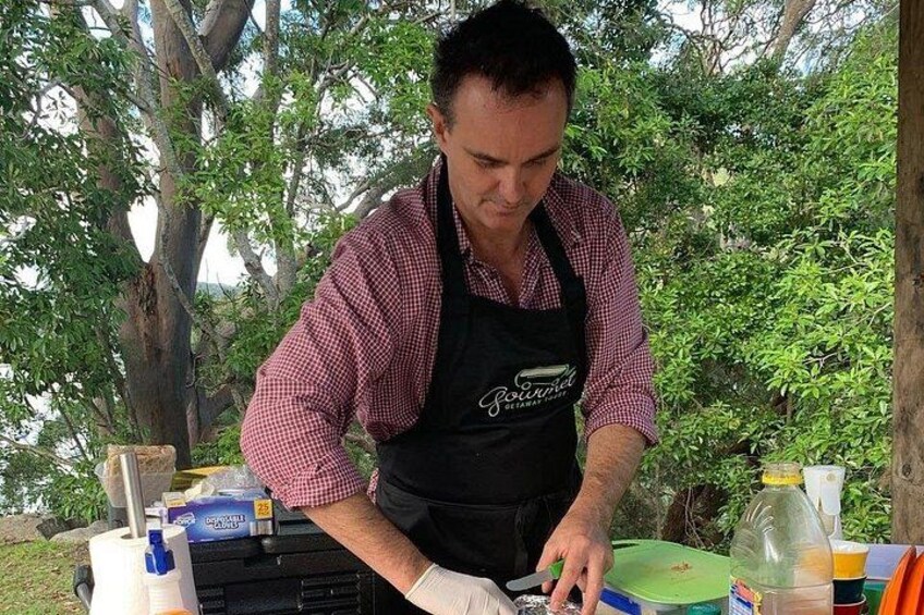 A person wearing a checkered shirt and apron, preparing food outdoors with a grill and various cooking utensils. 2