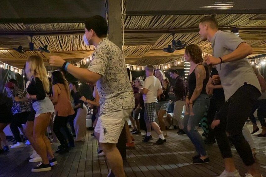 Människor dansar på en trångt belägen utomhusplats med trägolv och ett halmtak. 4