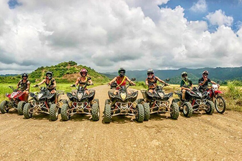 Five people on quad bikes on a dirt road with a mountainous landscape in the background. 3